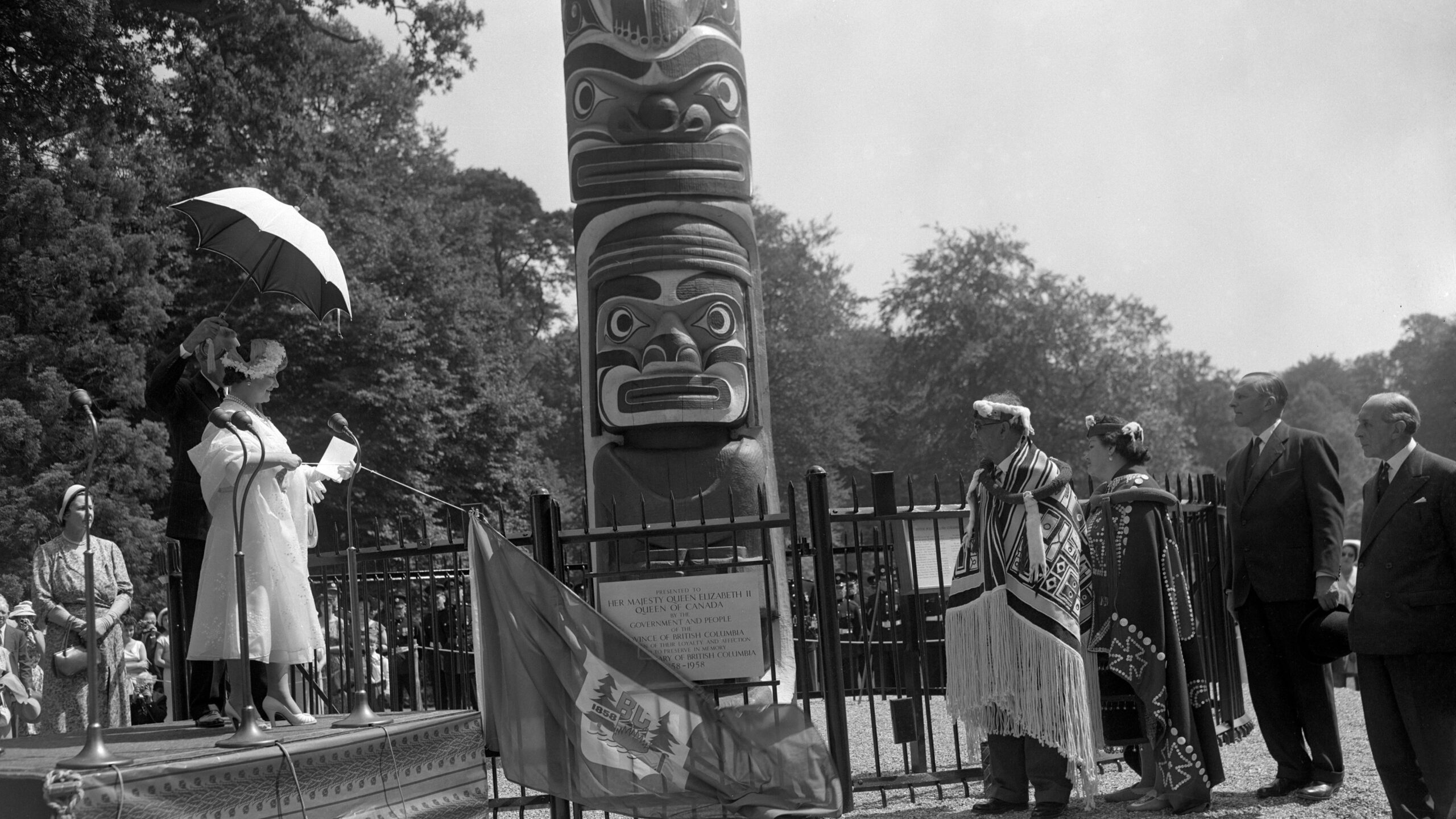 Queen Elizabeth The Queen Mother, who was deputising for Queen Elizabeth II, unveils the commemorative plaque at the base of the 100-foot totem pole, which has been erected in Windsor Great Park as a centenary gift from British Columbia. Watching are Chief Mungo Martin, of the Kwakiutl Indians, a tribe located on Vancouver Island, and his granddaughter, Helen Hunt. Chief Mungo Martin carved the elaborate ornamentation on the 12-ton pole.