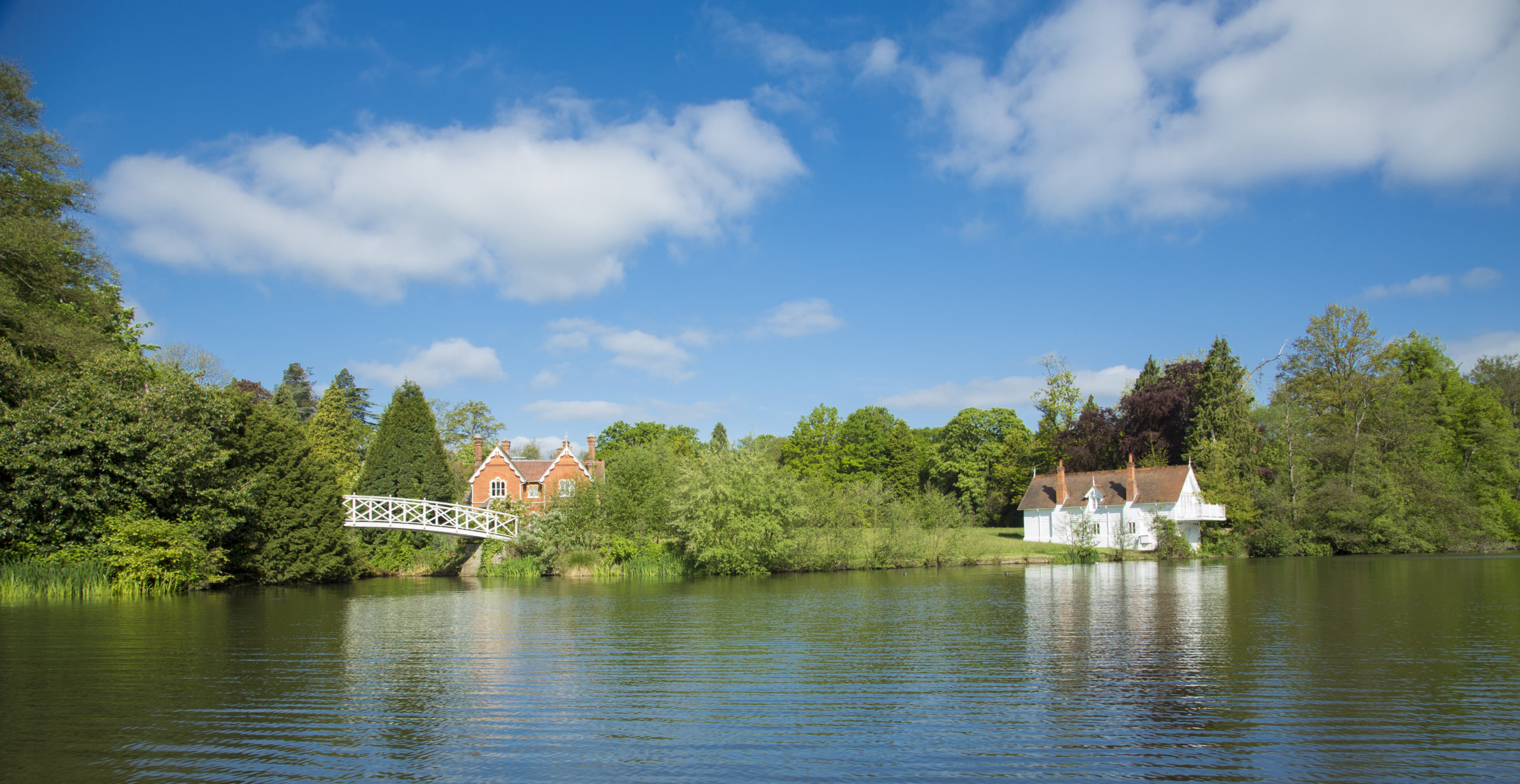 A Boathouse and property across a lake with a backdrop of trees and sunny skies 