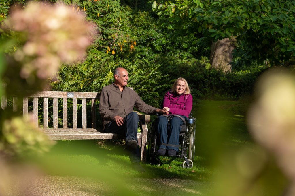 Two adults smiling in The Savill Garden. One sitting on a bench, the other in a wheelchair.