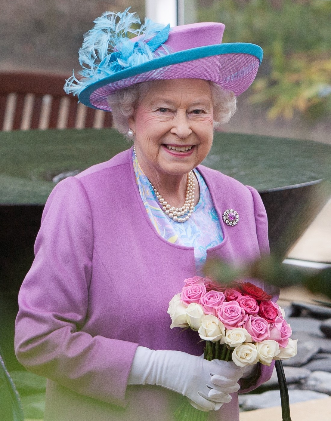 A photograph of Queen Elizabeth II visiting The Temperate House at The Savill Garden. She is smiling and holding a bunch of roses.