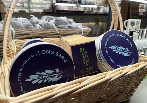 Rows of circular tubs with blue and purple lids in a wicker basket with a row of brown boxes. All are branded with an image of lavender.
