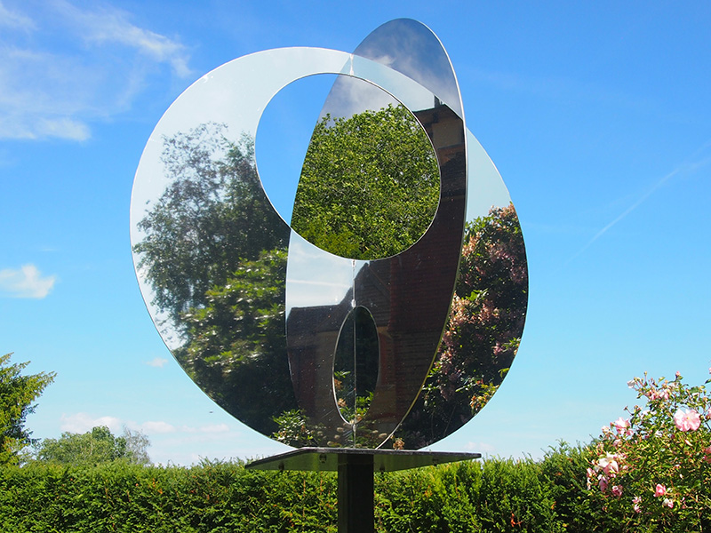 A sculpture of two circular mirrors that intersect in the middle. Behind is a below sky with a green hedge.