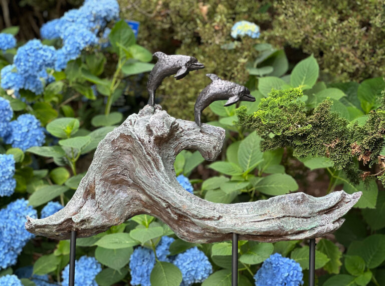 A sculpture of two metal dolphins leaping from a wave made of wood attached to two poles staked to the ground. In the background is a hydrangea with blue flowers and green leaves.
