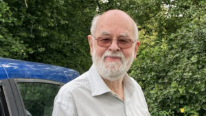 A man stands in front of a green hedge, wearing a check shirt and glasses. He has a grey beard and moustache and a bald crown.