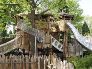 A picture of two wooden trees houses. The one on the right has a metal slide trailing down it, while the treehouse on the left has a metal climbing tube that extends out of the treehouse and wraps around a tree.