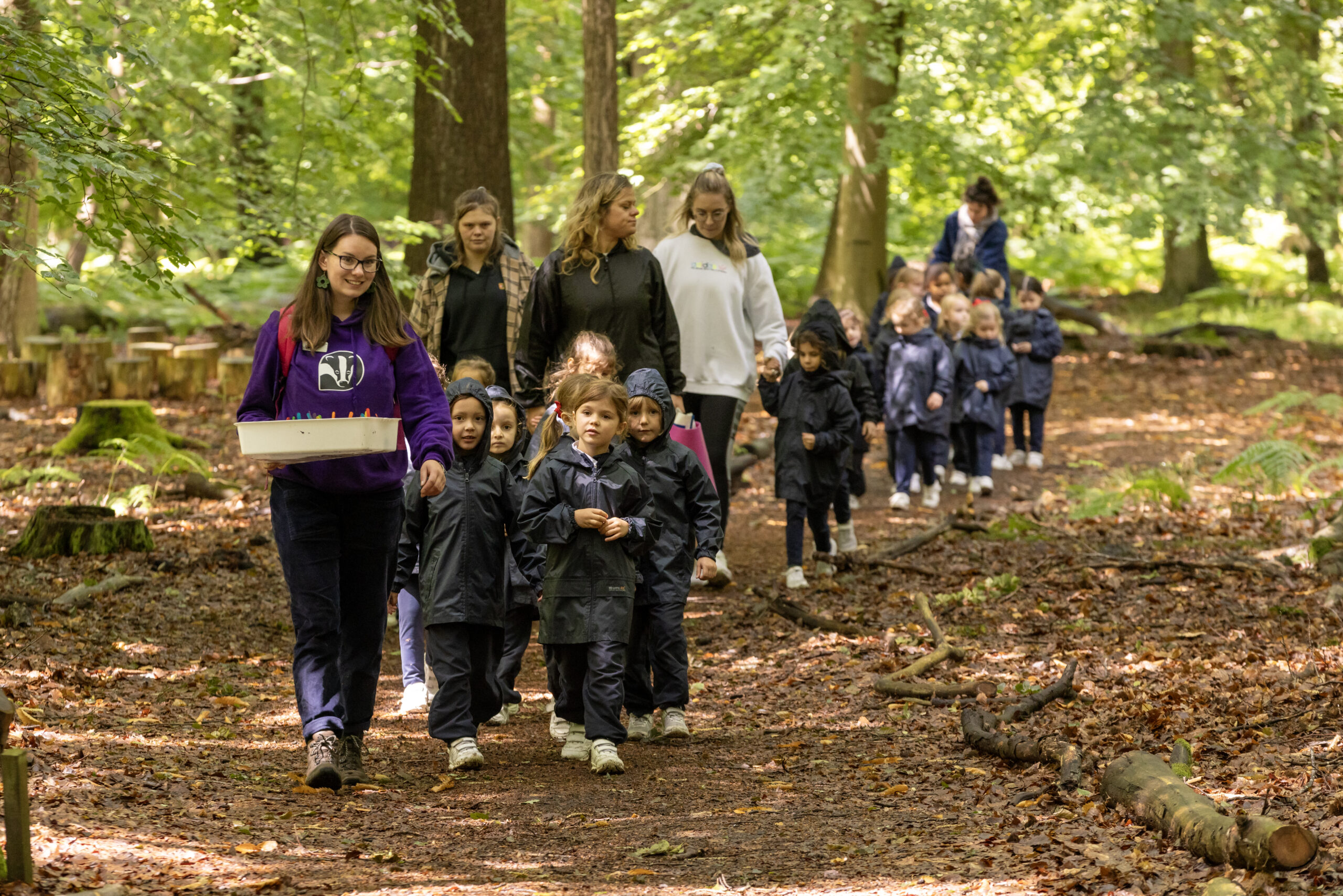 A group of children being led and accompanied by some adults on a bark chipped path in the forest.