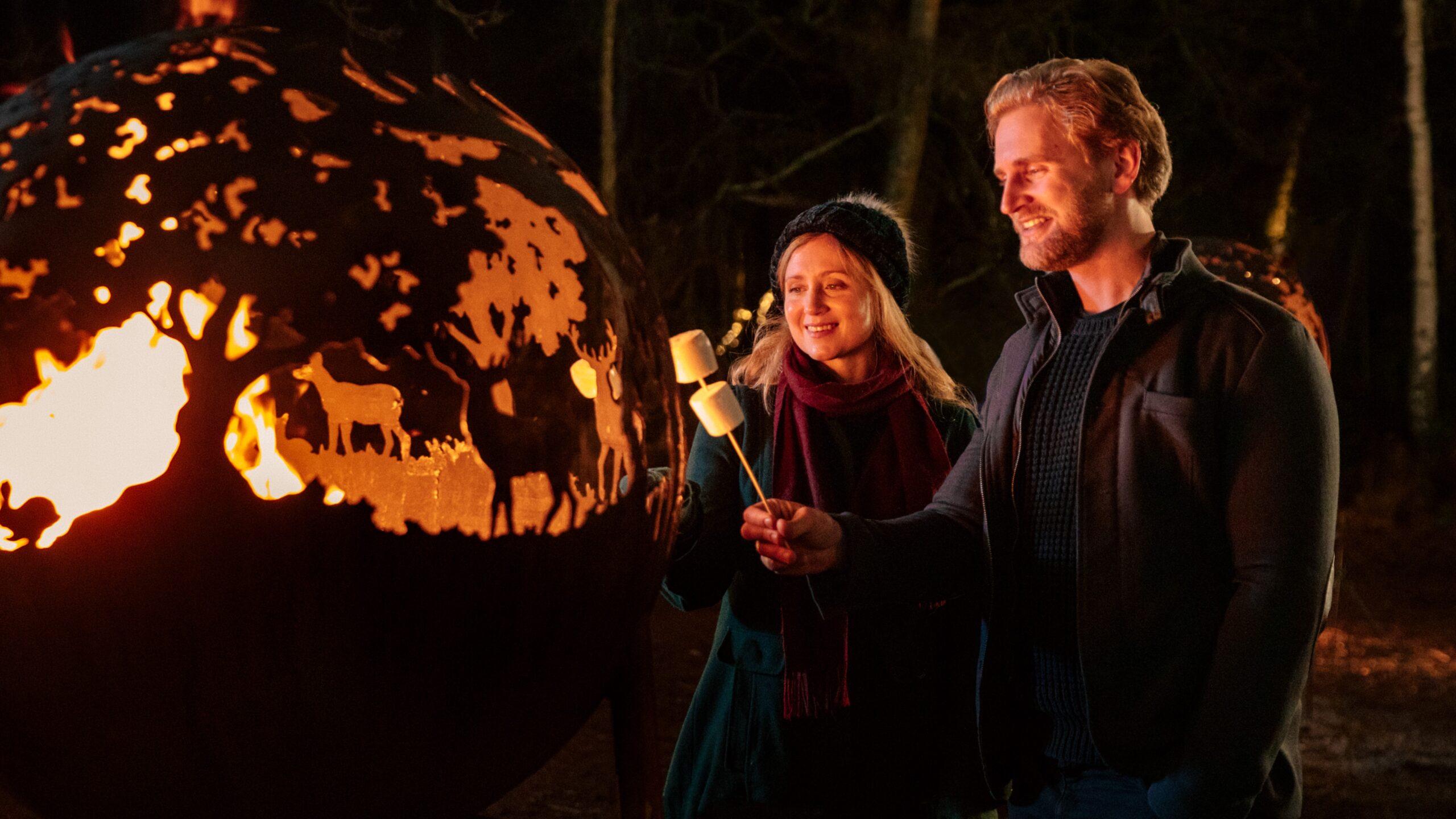 Adults toasting marshmallows in front of an open fire which is lit within a steel framed globe.