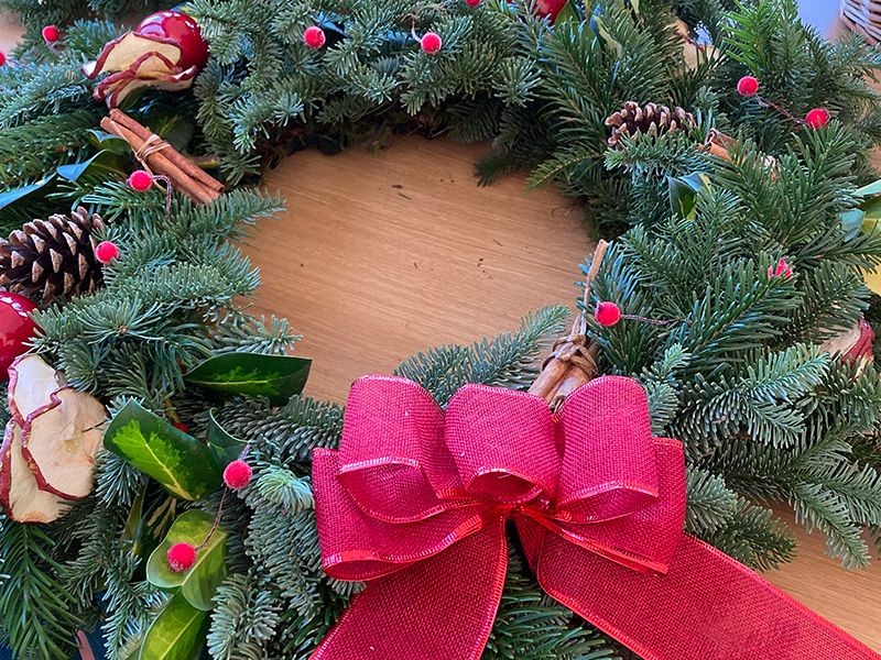 A Christmas wreath of fir leaves, pine cones, dried fruit, cinnamon sticks and a red ribbon.