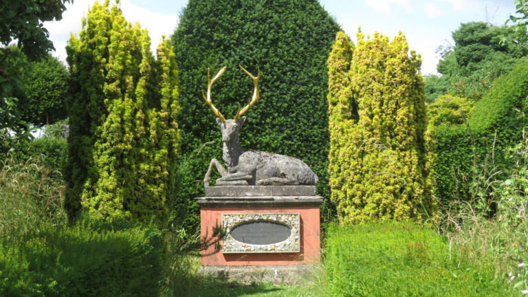 A statue of a deer with gold antlers sits on a pedestal. Behind it is a tall green hedge with tall green grass beside it.