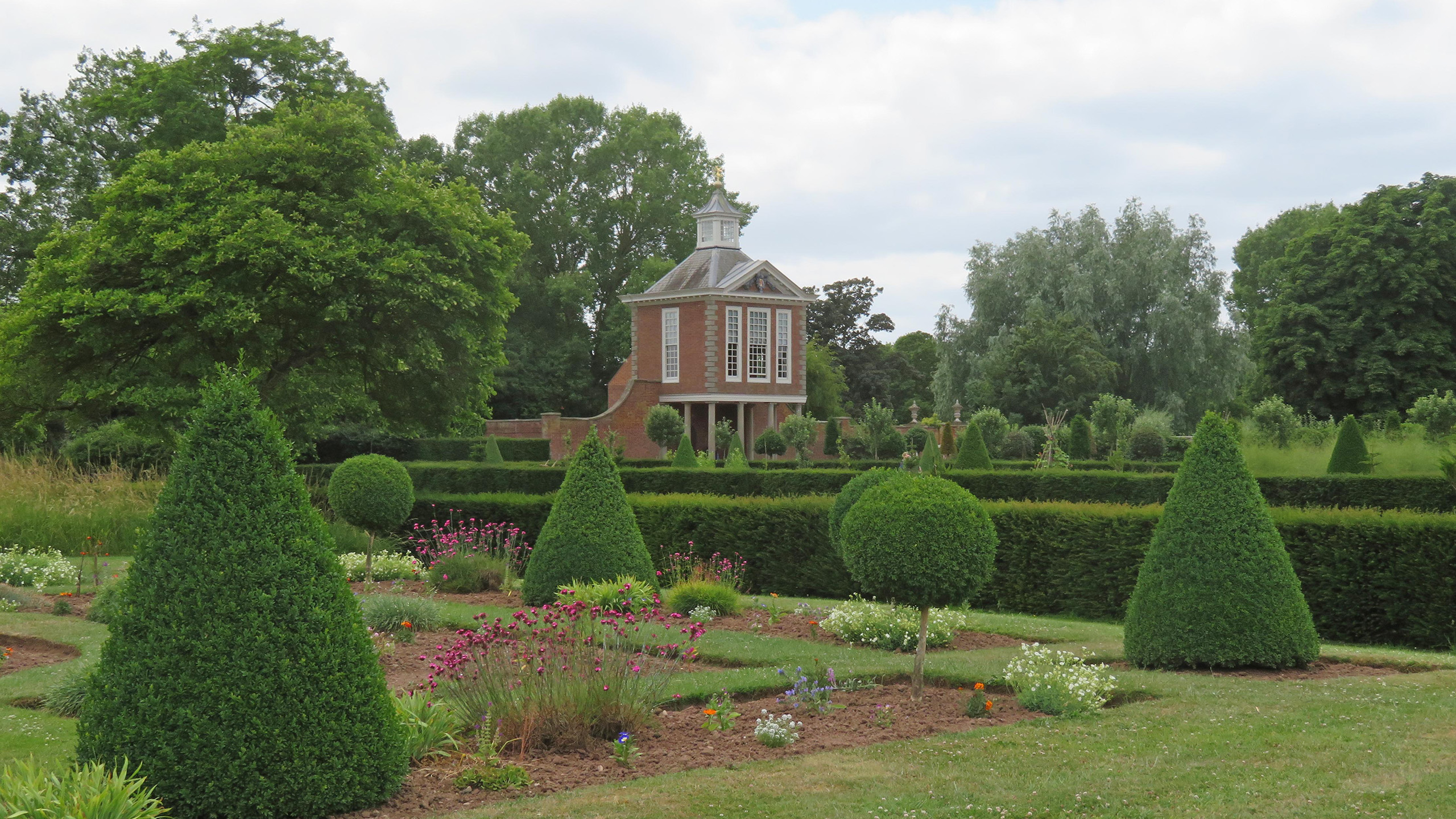 A formal garden of yew hedges shaped in cones and balls, with hedges dividing the further gardens in the distance. A brick building stands in the background beside tall green trees.