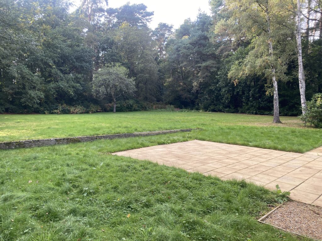 View of the grass lawn and patio surrounded by tall trees. Pinewood Lodge, Swinley Road, Ascot, SL5 8BA