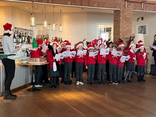 A row of school children in red jumpers and grey trousers stand inside holding sheet music and sing towards the camera. Behind them is a brick pillar and arches. 