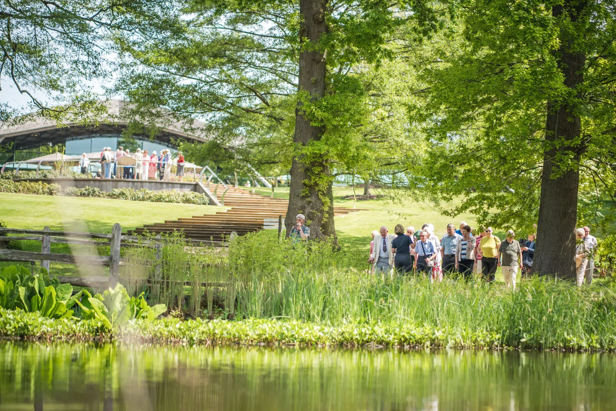 Group visiting The Savill Garden with views of the Visitor Centre, tress and pond.