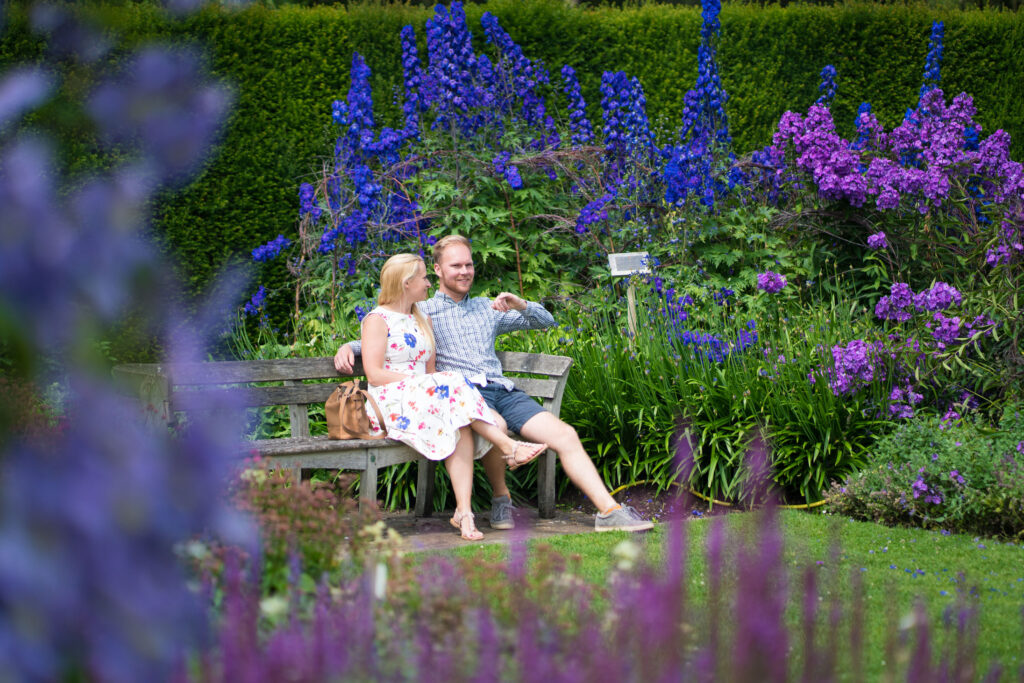 Two adults sitting on a bench in The Savill Garden surrounded by purple and pink flowers.