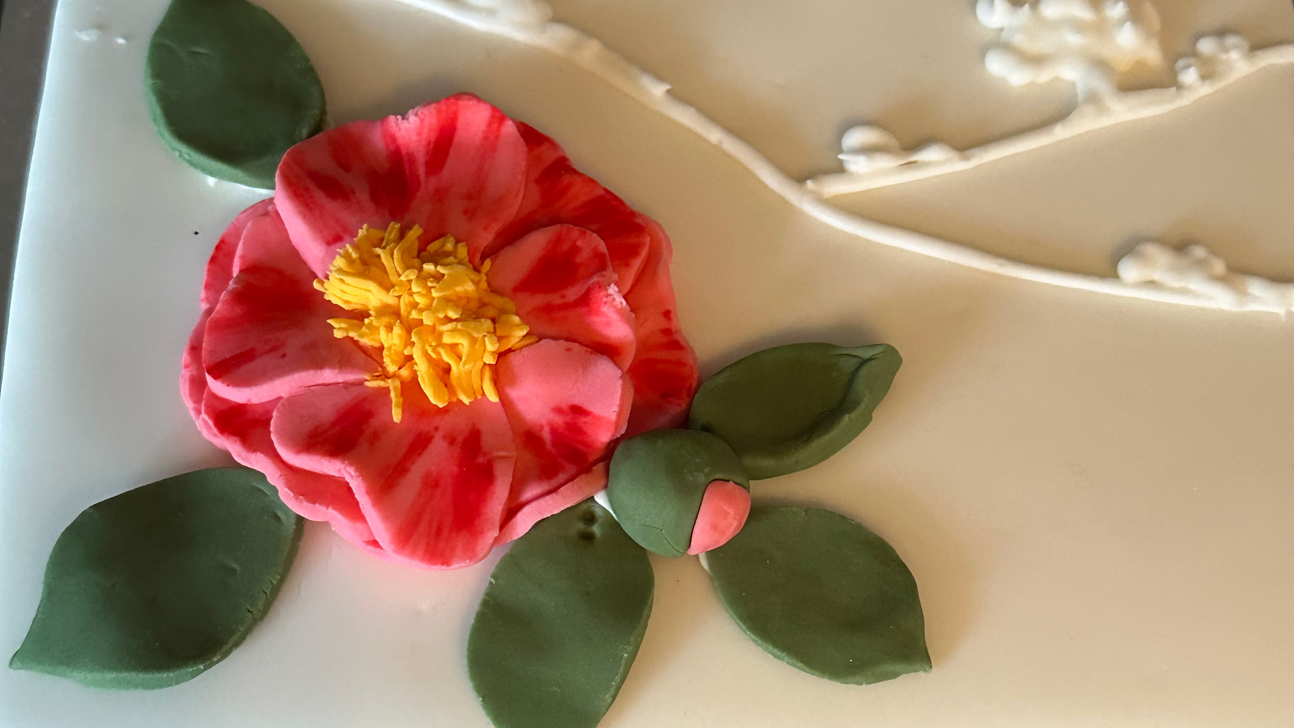 A close up of a red camellia flower and green leaves made of fondant icing on a white cake.