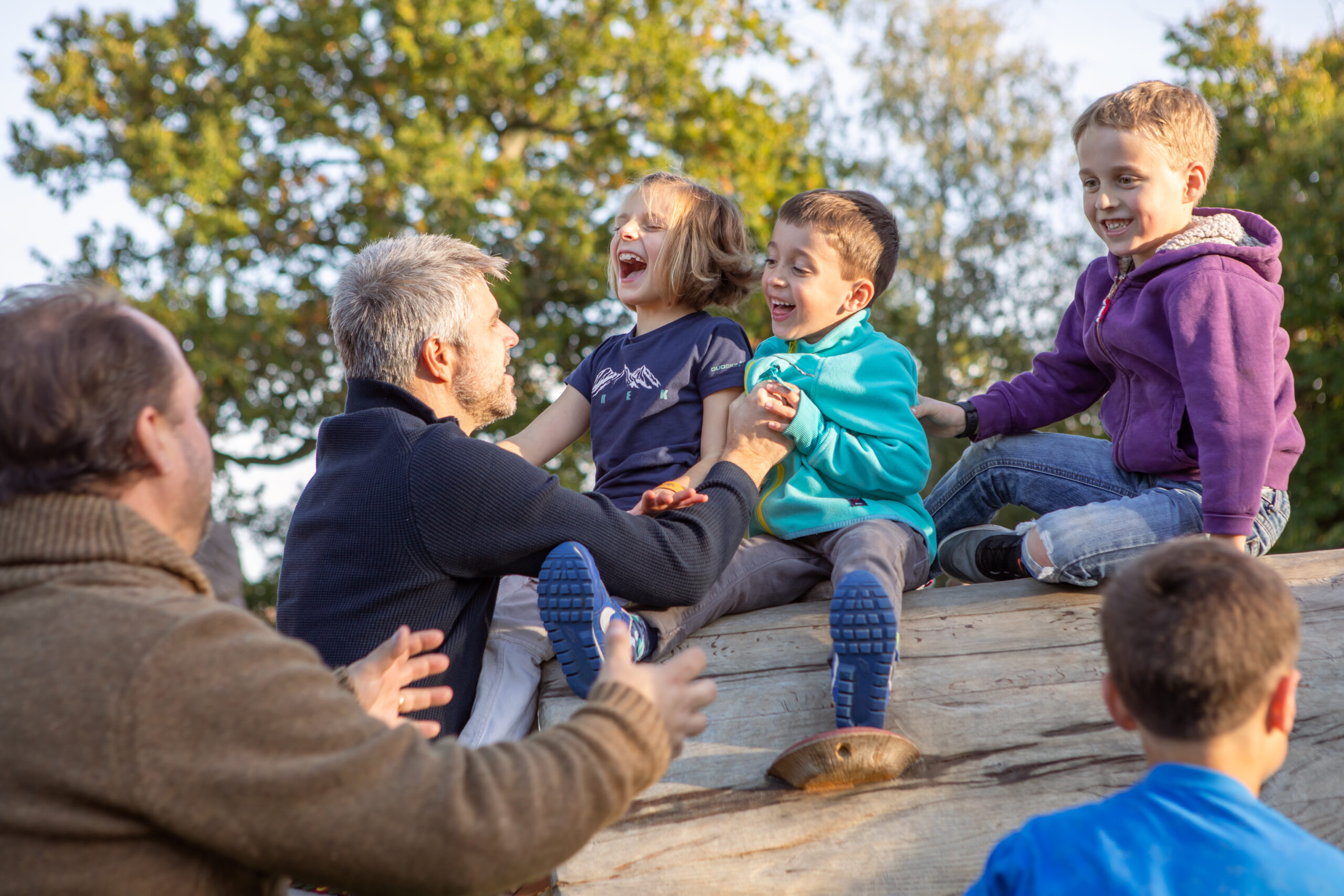 Three children sitting on a tree trunk, smiling. Supervised by two adults.