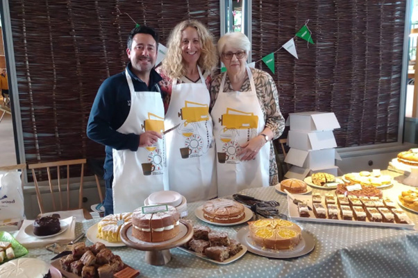 Three people wearing aprons standing behind a table laid with cakes.