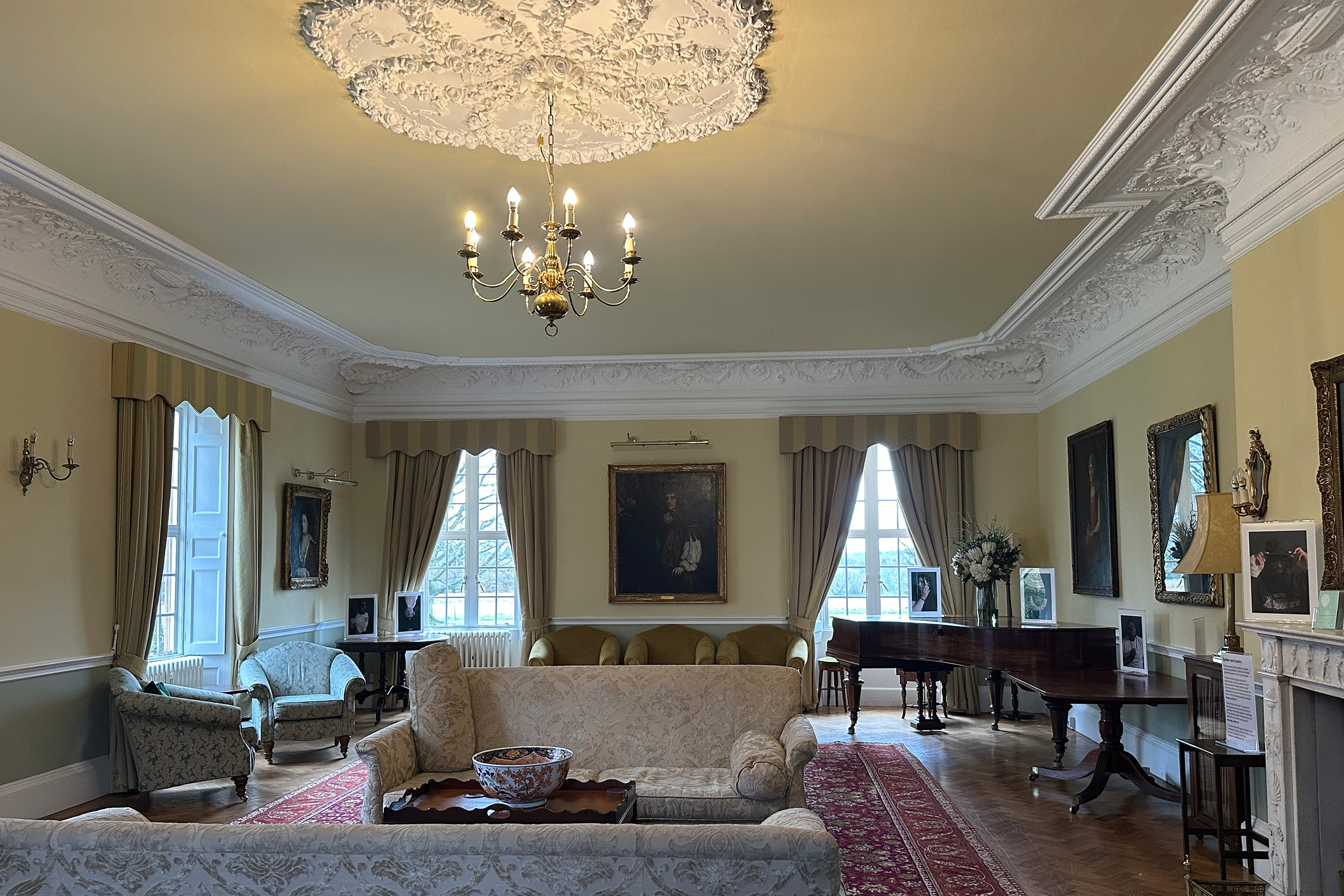 A Drawing Room with upholstered seating, a large piano, a fireplace and plastered ceiling.