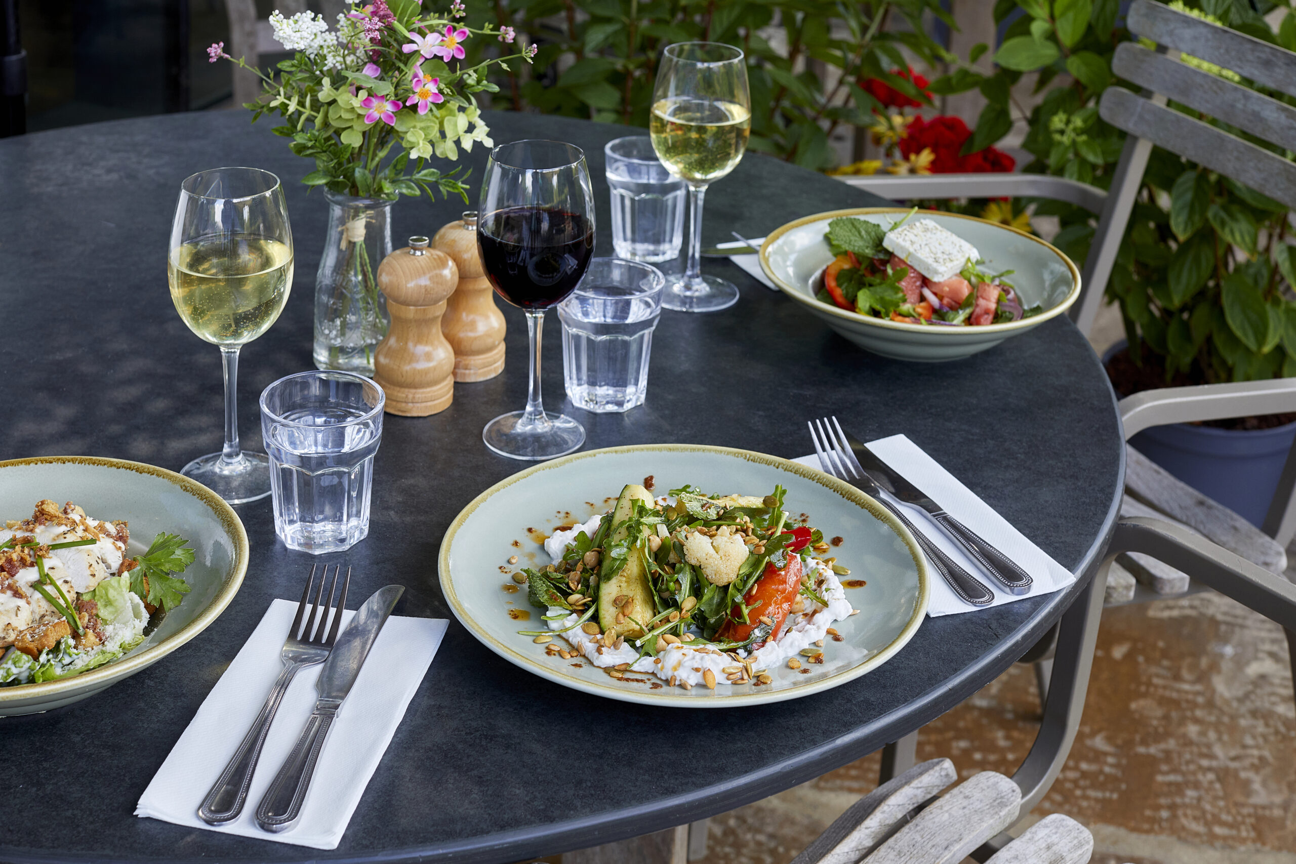 Outdoor table laid with food, drinks and a small vase of flowers.