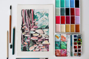 A watercolour painting of a tree and fungi beside a palette of paints and paintbrushes.