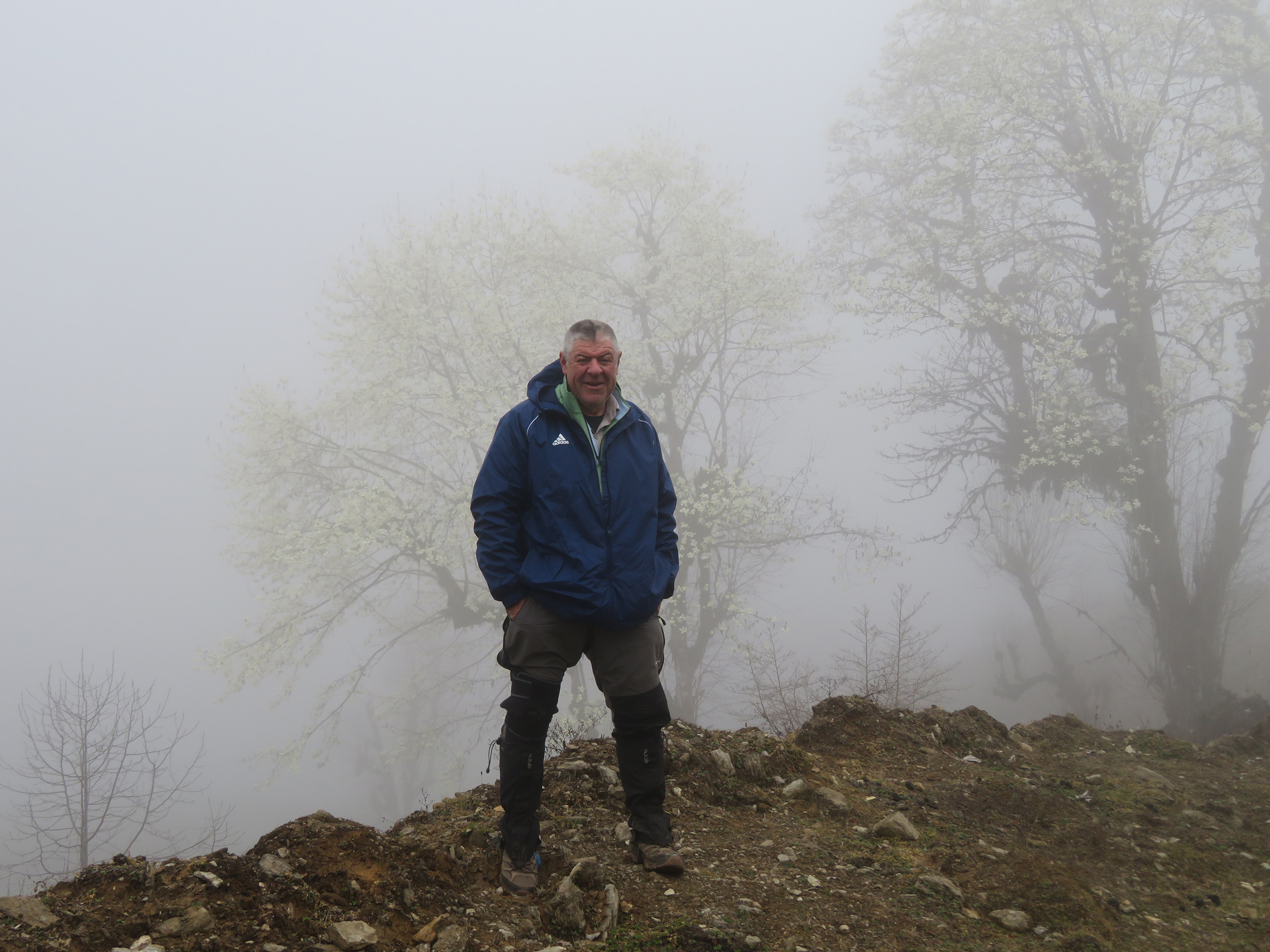 A person wearing clothing for a cold climate standing on a hill with trees and mist behind.