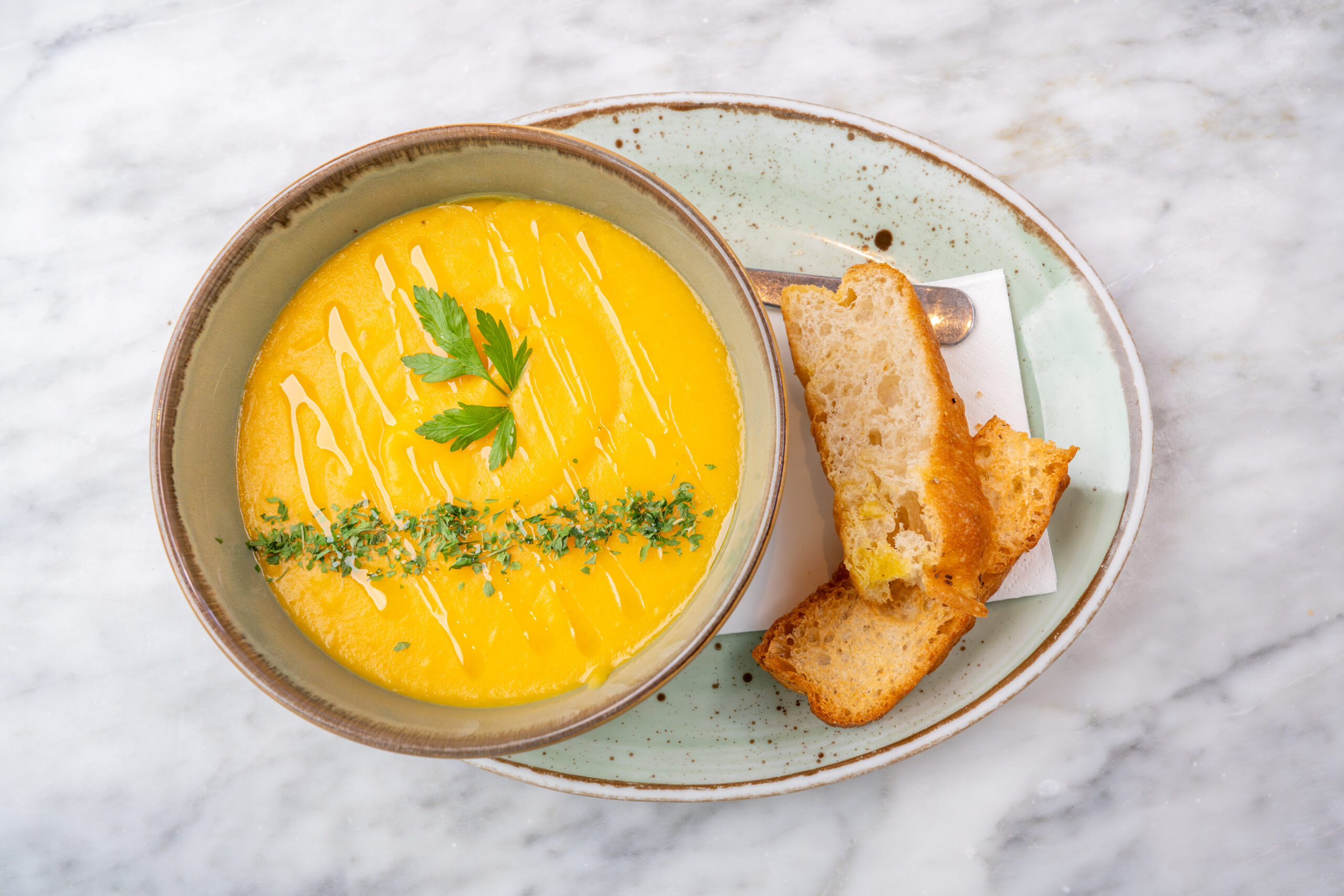 A bowl of butternut squash soup served on a plate beside fingers of crusty bread.