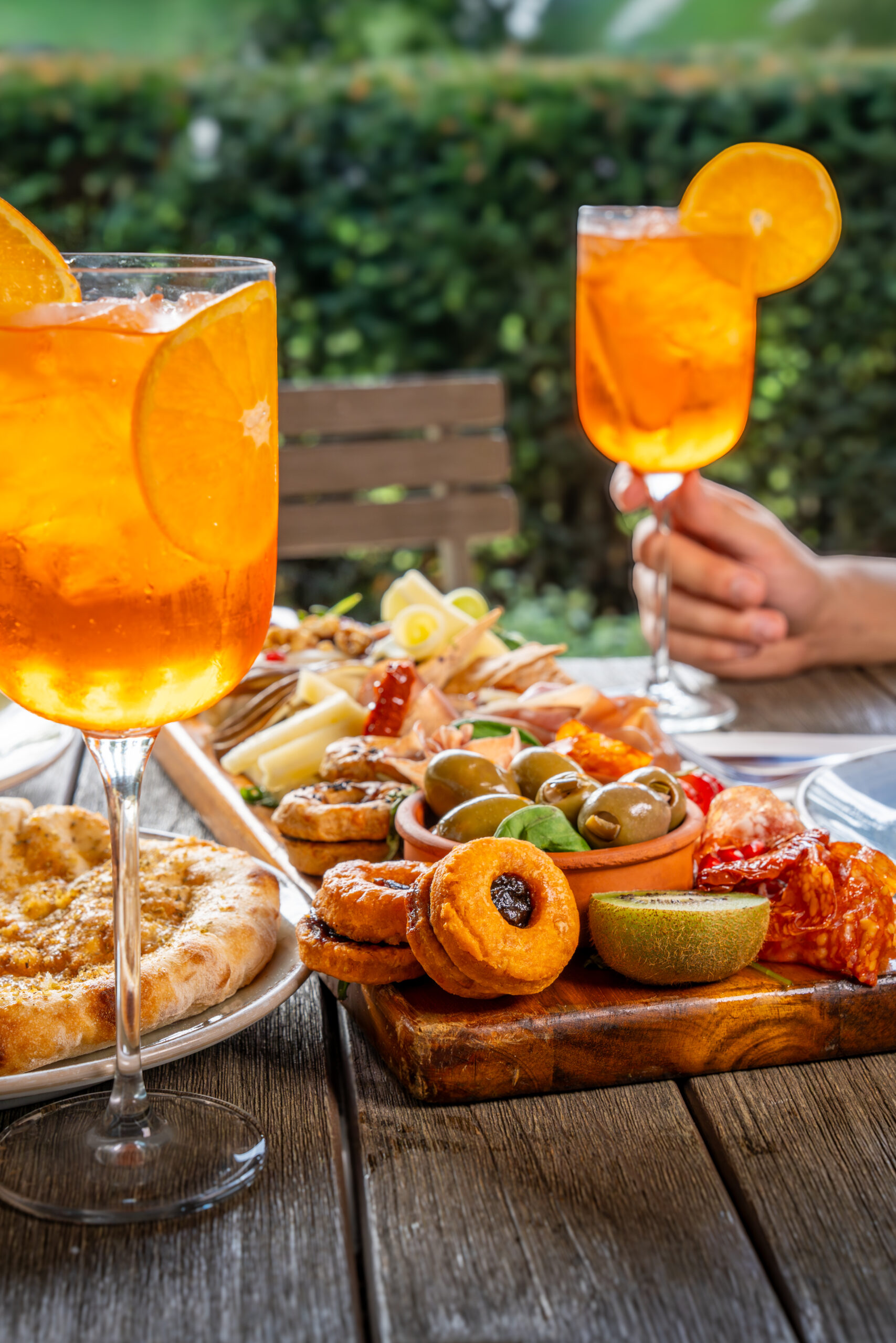 A wooden board of antipasto served on a table with two glasses of aperol spritz/