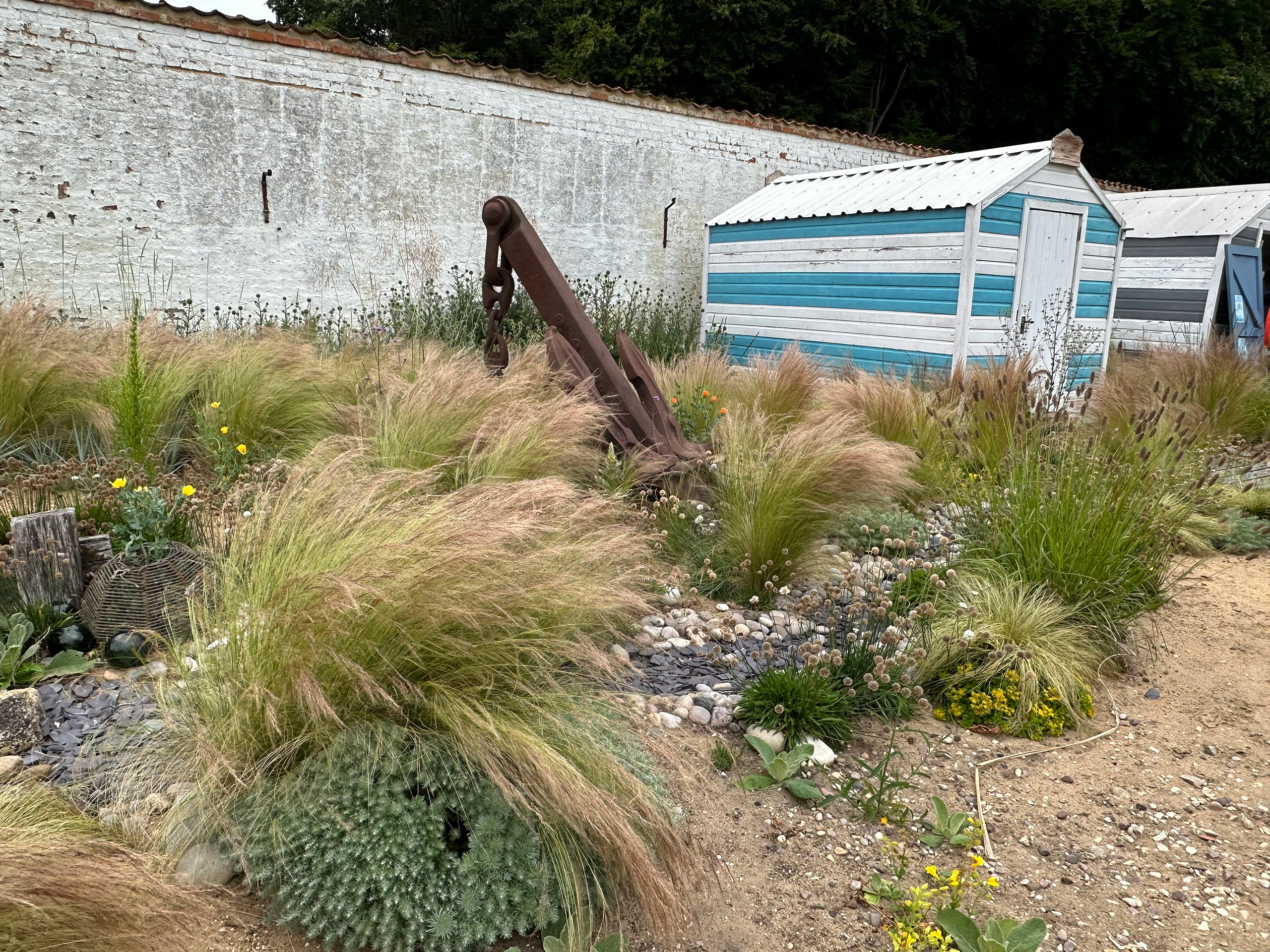 A large ship anchor sits amongst grasses. Further in the background are beach huts and sand.