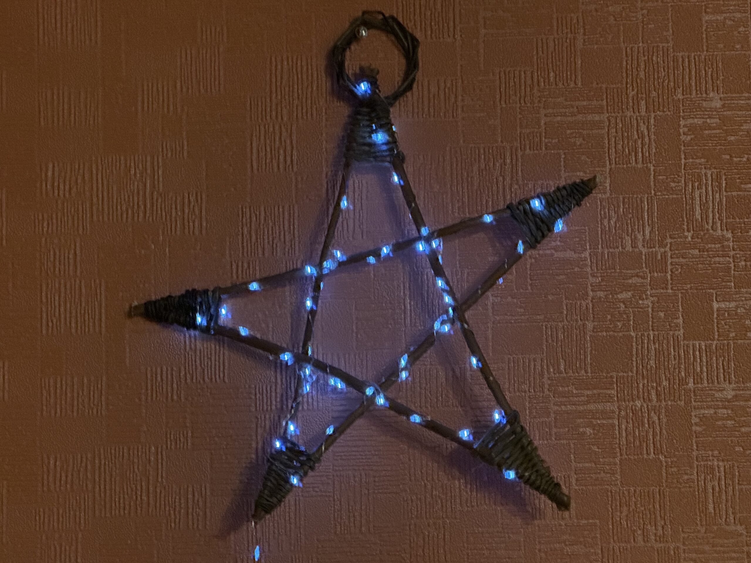 A finished willow star hanging on a textured wall, wrapped with small blue LED lights that illuminate the outline of the star shape.