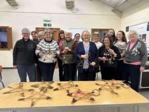 A group of people standing together in a community hall, each holding a star-shaped craft made from willow sticks. A table in front of them displays several completed willow stars. The room has cream-colored walls, wooden doors, and fluorescent lighting.