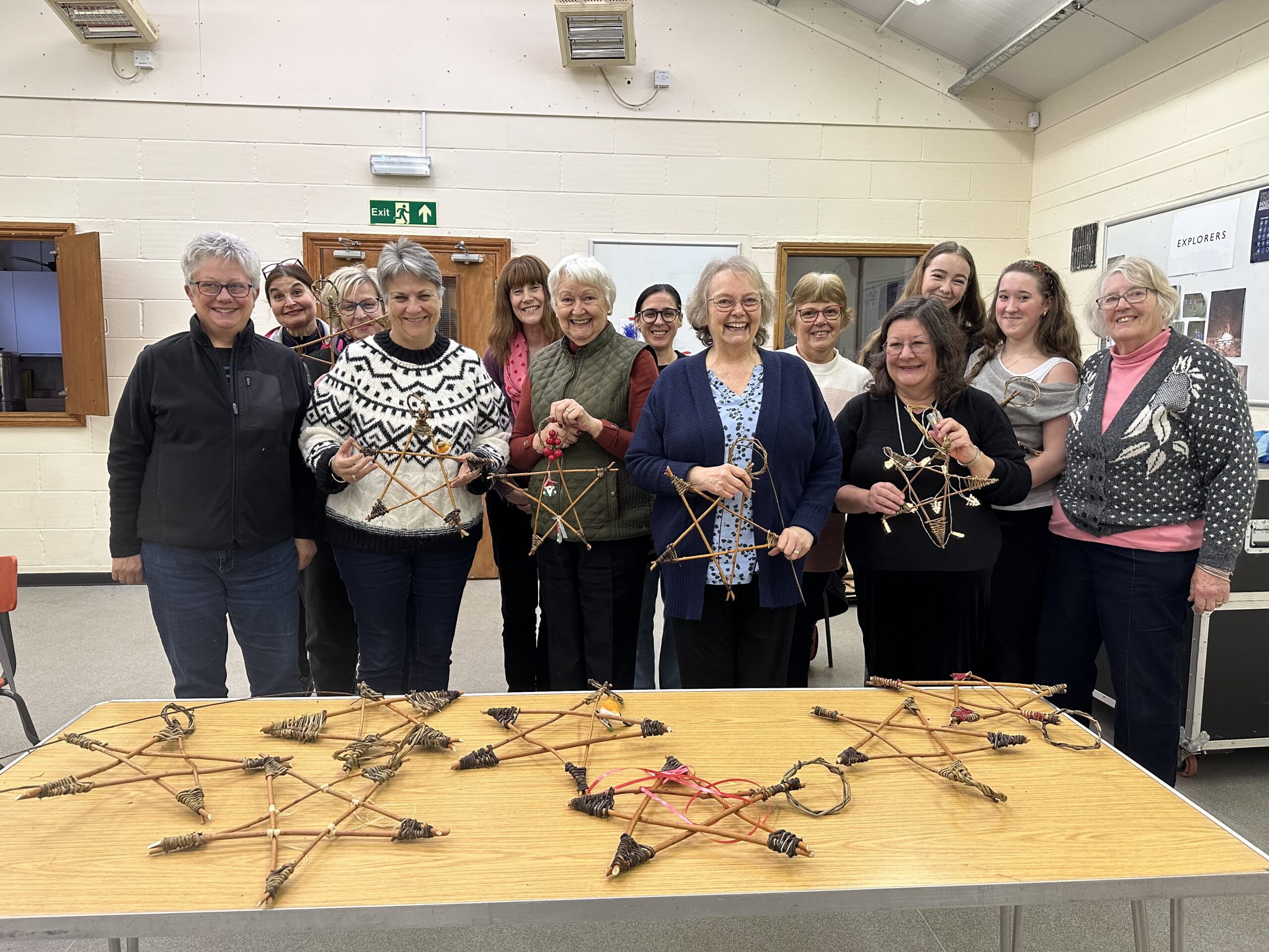 A group of people standing together in a community hall, each holding a star-shaped craft made from willow sticks. A table in front of them displays several completed willow stars. The room has cream-colored walls, wooden doors, and fluorescent lighting.