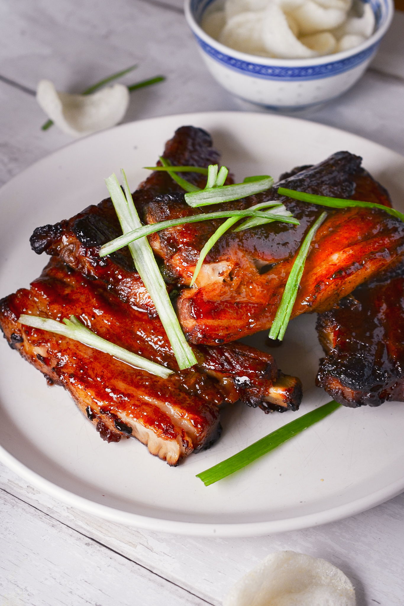 Char Siu Pork Ribs garnished with sliced Spring Onions alongside a bowl of Prawn Crackers.