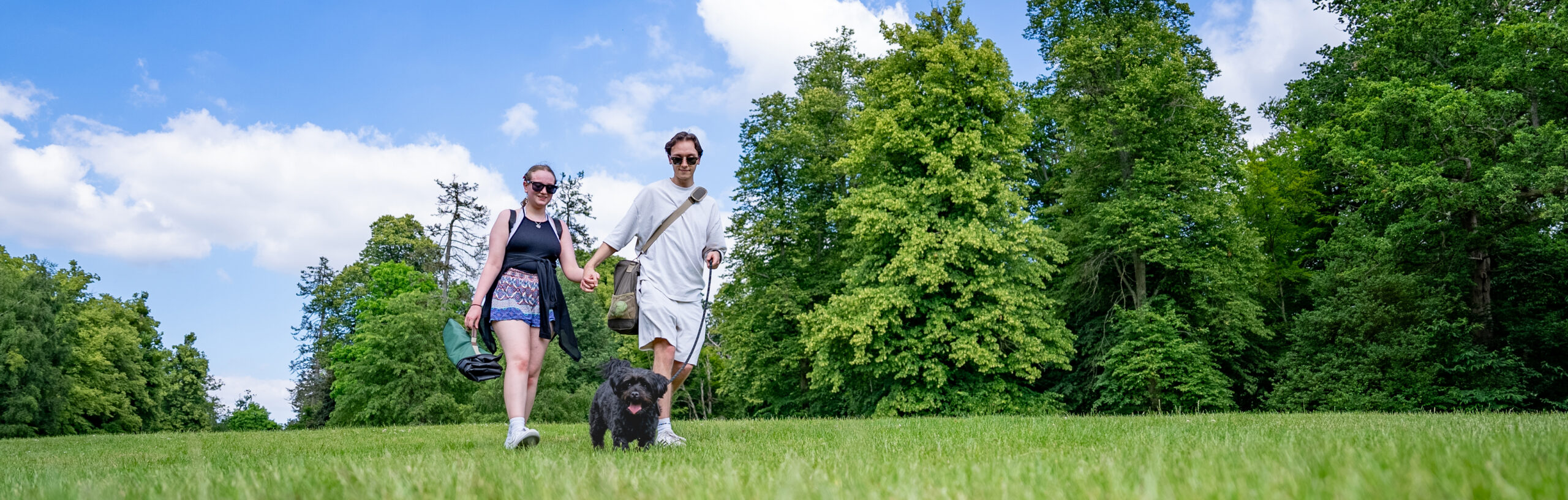 Two visitors walk with a dog on a lead along a grassy lawn. They are carrying a rucksack and picnic blanket.