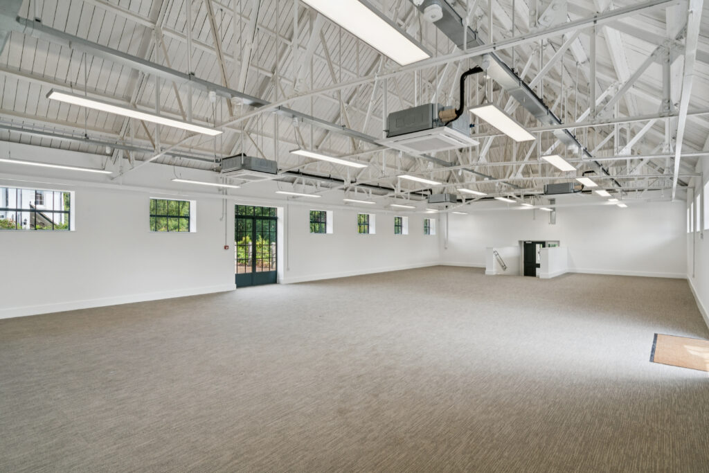 Wide interior view of a bright event or meeting space with exposed white roof trusses, large windows, and open floor area.