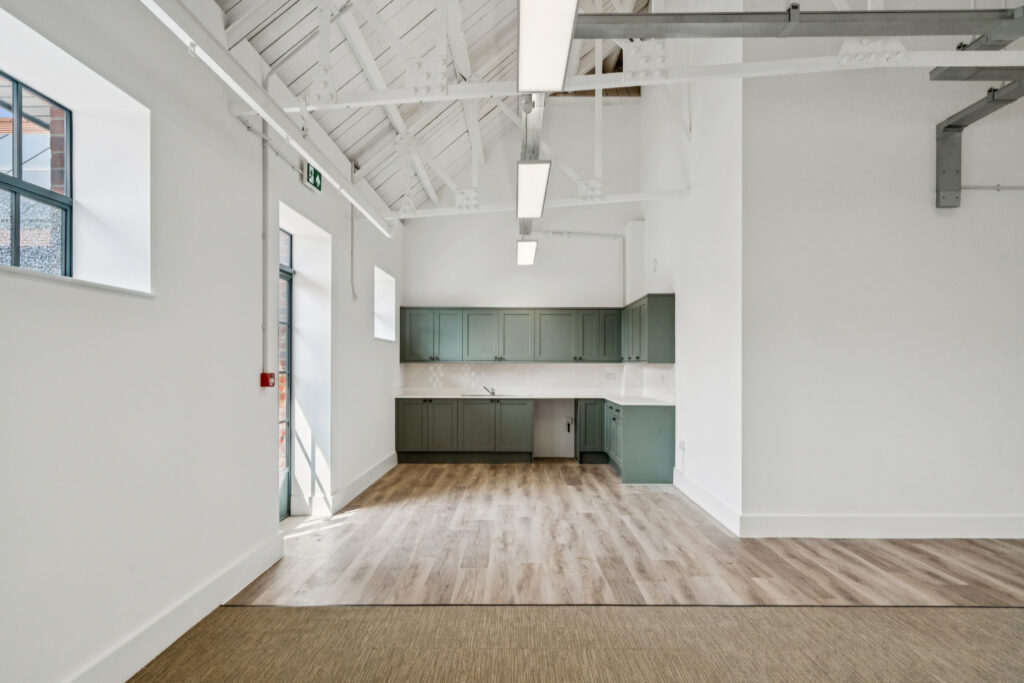 Kitchen area with sage-green cabinets, white worktops, wood-effect flooring, and high ceilings with exposed painted beams.
