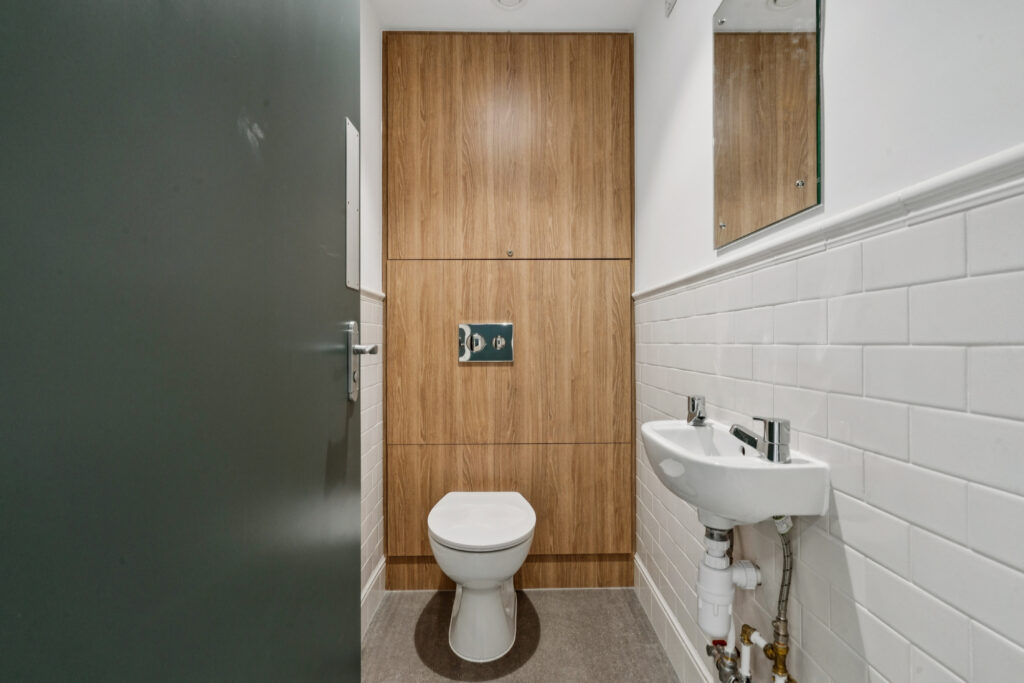 Accessible washroom with a toilet, wall-mounted sink, white tiled walls, wood-effect panelling, and a mirror.