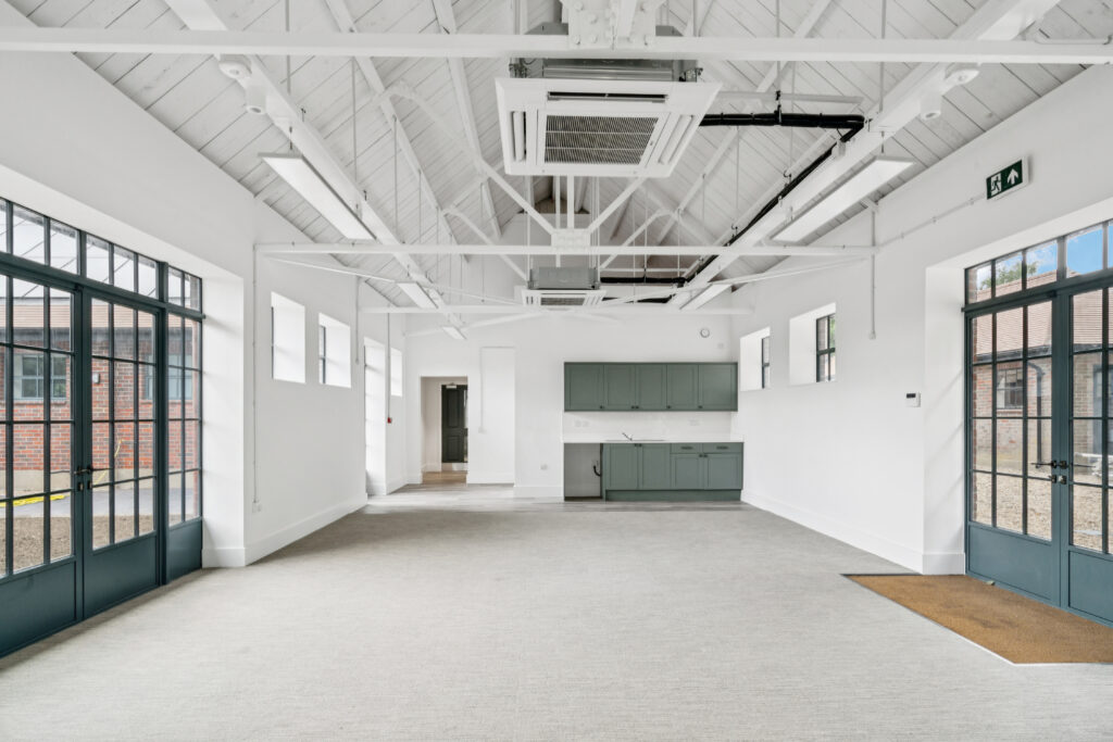 Open-function room with high vaulted ceiling, large glazed doors, exposed white roof trusses, and a kitchenette at the far end.