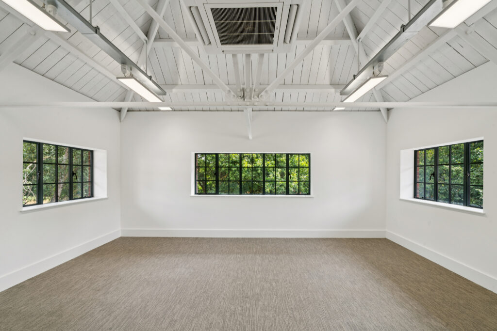 Empty, bright room with white walls, exposed vaulted timber ceiling, suspended lighting, and three large black-framed windows overlooking trees.