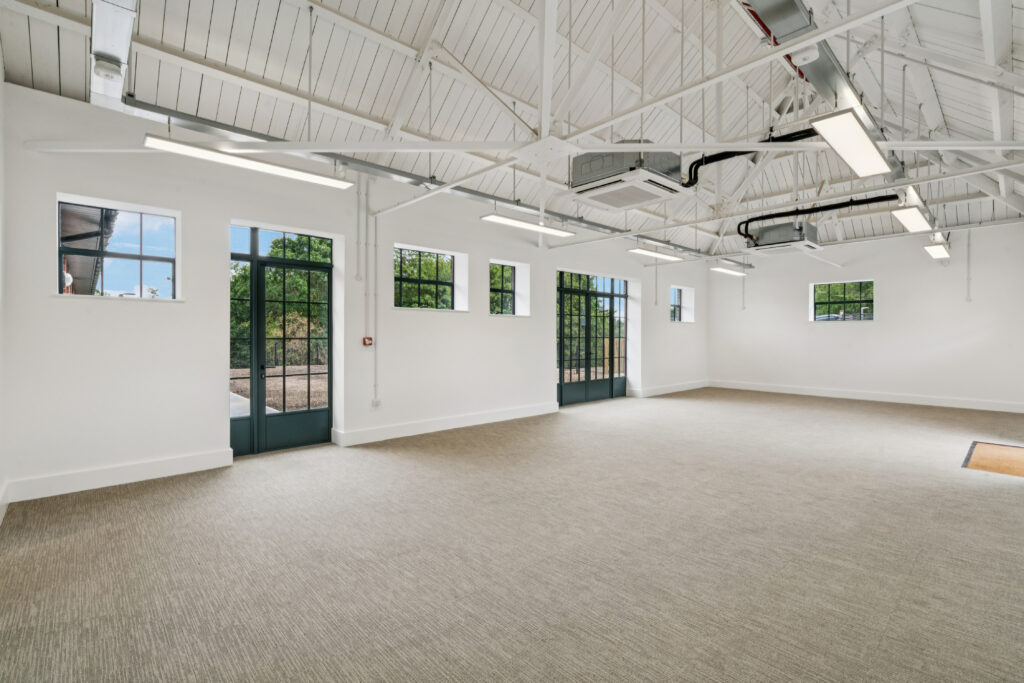 Spacious open-plan hall with white vaulted ceiling, exposed beams, large glazed doors and windows, and views of surrounding greenery.