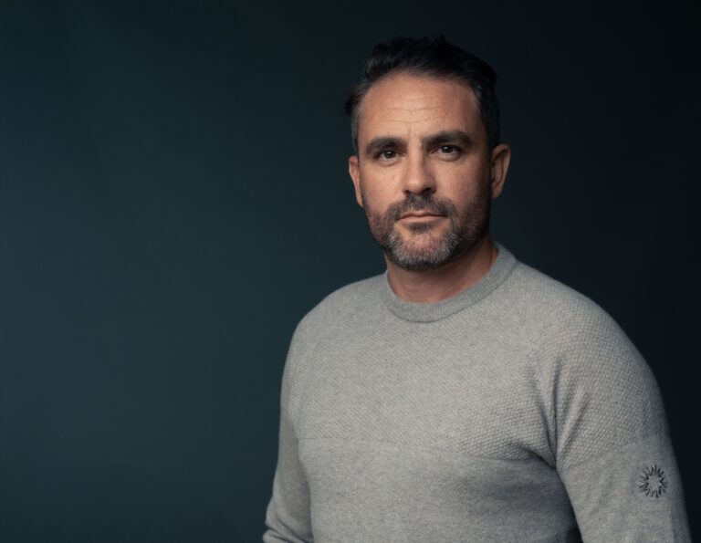 Portrait of a person wearing a light grey knitted jumper, standing against a dark blue studio backdrop. The image is framed from the waist up, with the person facing the camera and positioned slightly to the right of the frame.