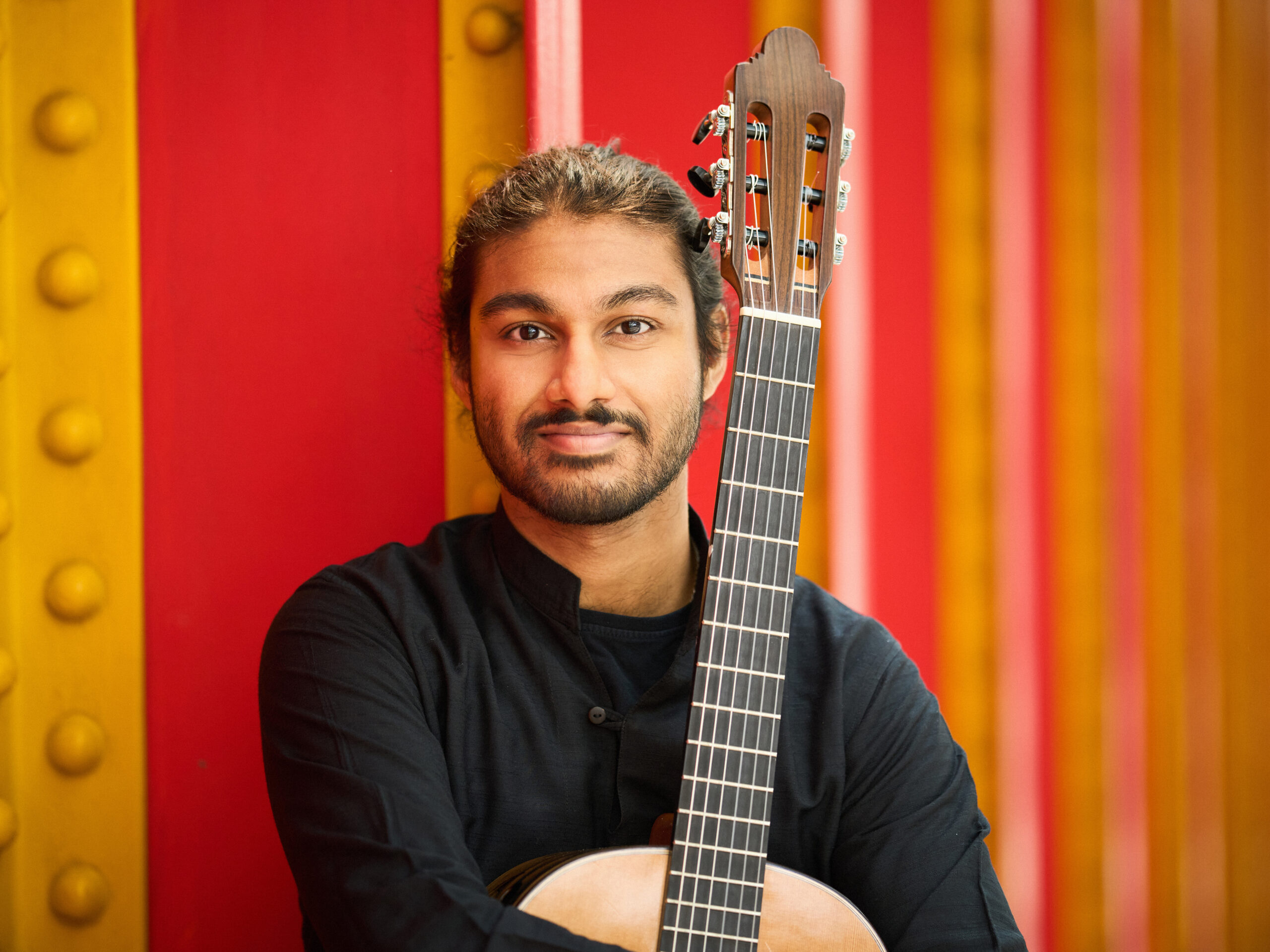 Portrait of a person holding a classical guitar, photographed against a colourful background of vertical red, orange and yellow panels. The person is wearing a dark long-sleeved top and is positioned in front of the guitar neck, with the image framed from the chest up.