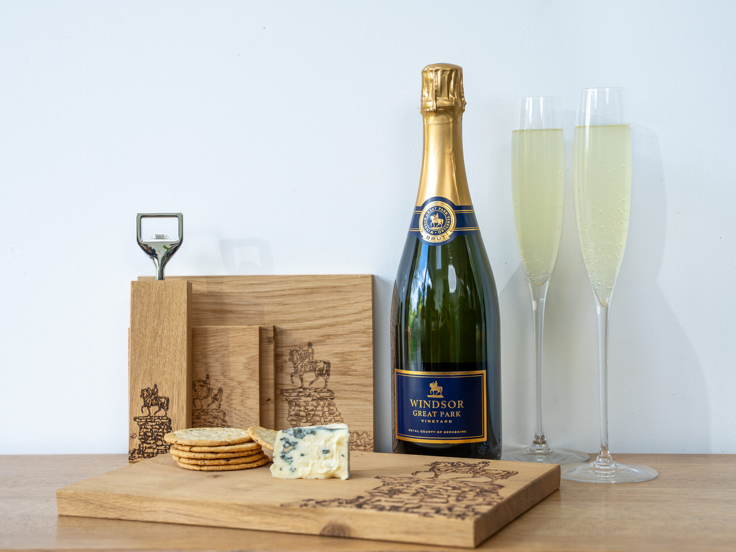 A bottle of Windsor Great Park Vintage Sparkling Wine with two glasses, a cheese board and wooden boards from Selbrae.